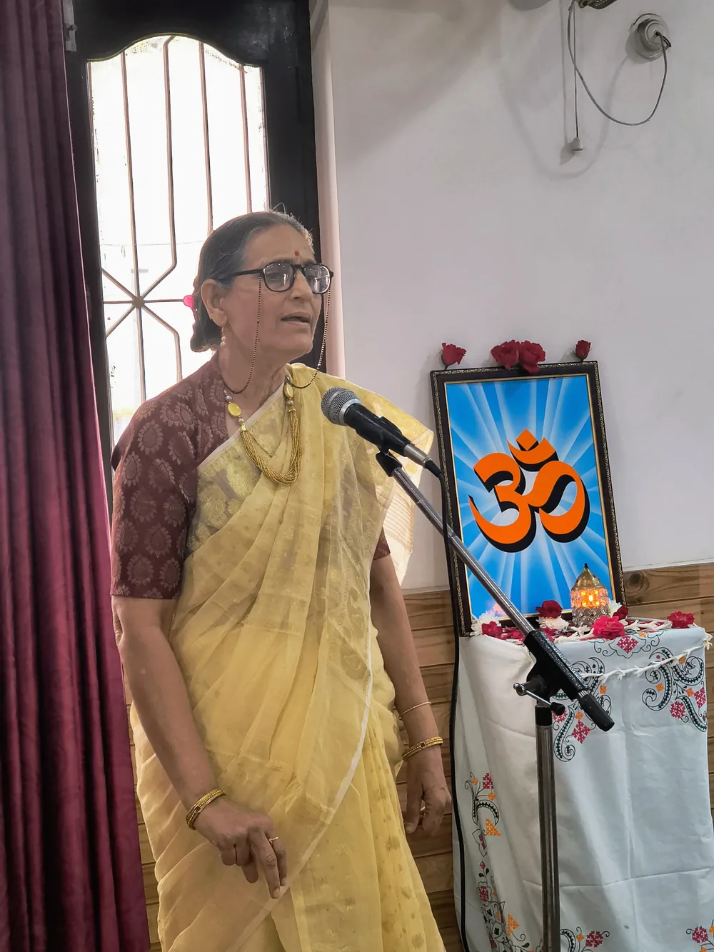 Sanjivani Joshi speaking at a microphone during a session, beside an Om emblem